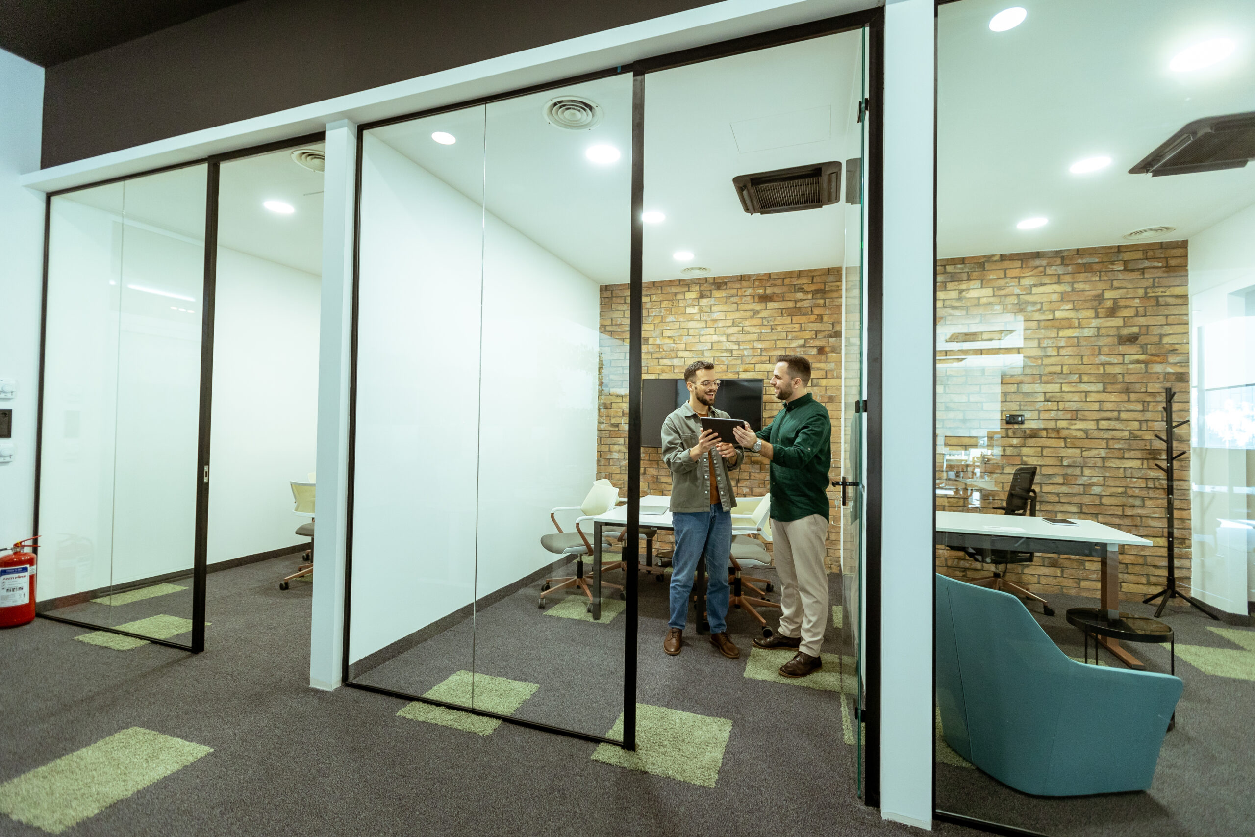 Сотрудники стоят и обсуждают документы в офисе с перегородками из стекла и кирпичной стеной
