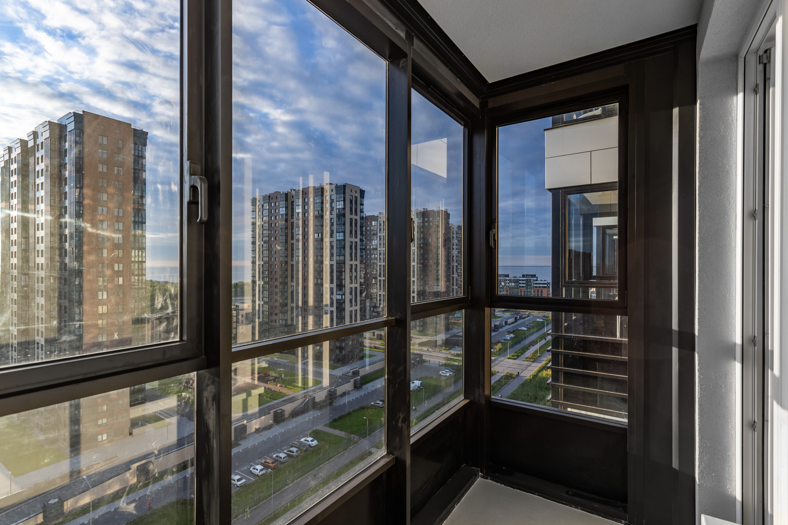 Застеклённый балкон с тёмным профилем и панорамными окнами, вид на современные жилые дома и улицу внизу.