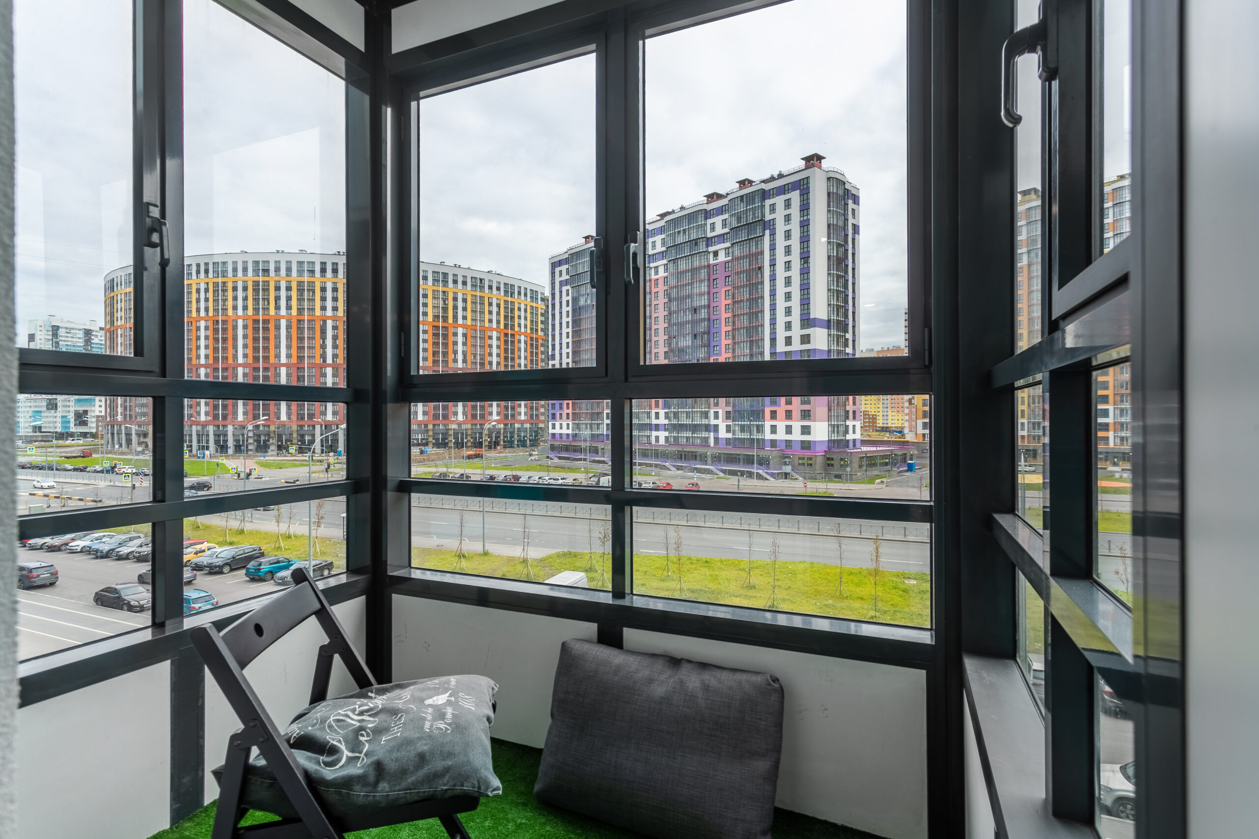 Застеклённый балкон с черным профилем и видом на современные жилые дома, внутри складной стул и подушка на полу.