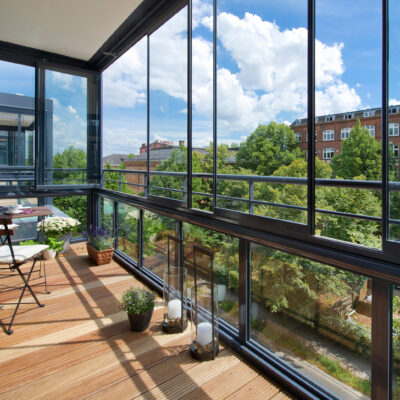 Остеклённый балкон с панорамными окнами, деревянным полом, мебелью и цветами, вид на городские дома и зелёные деревья.