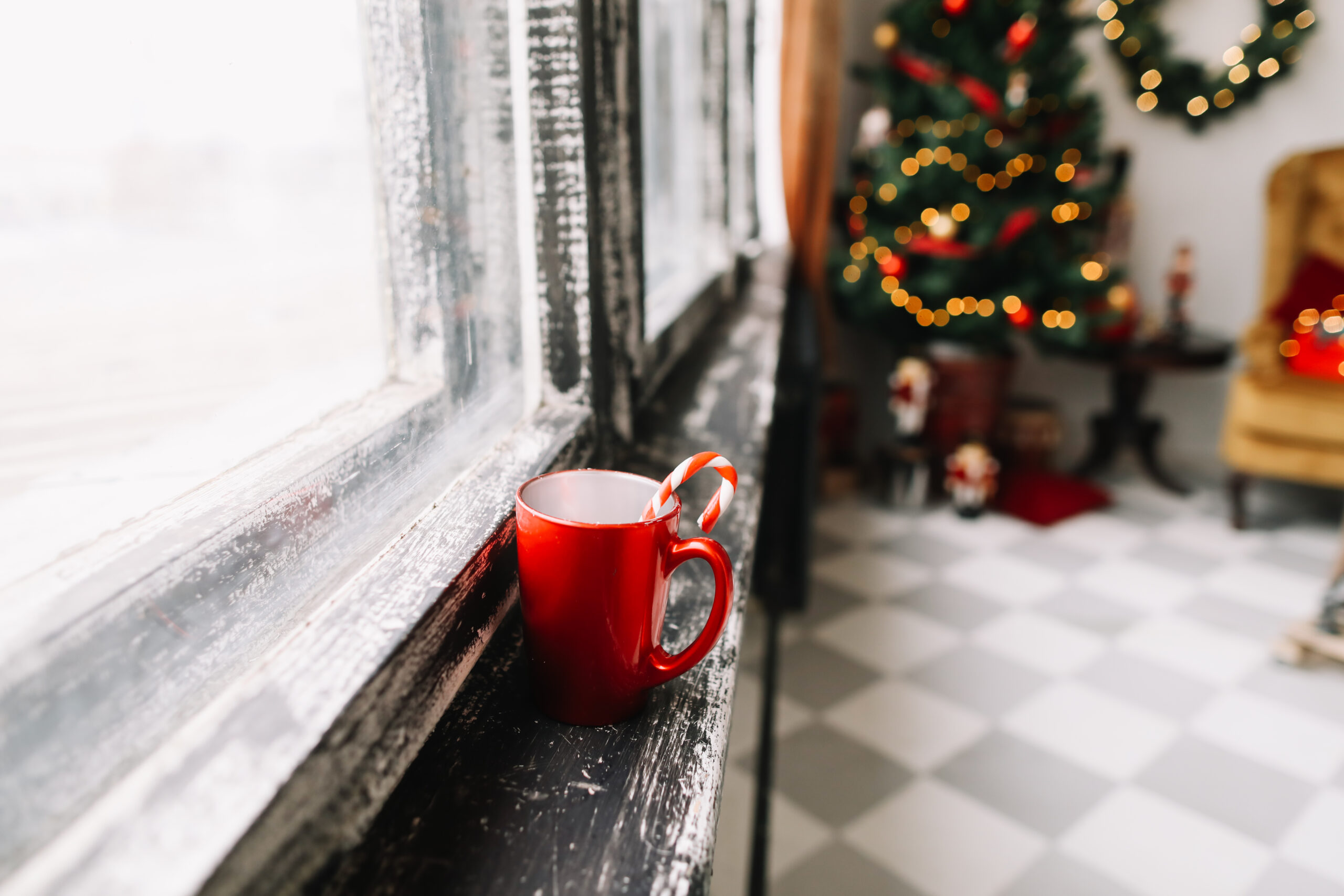 Красная кружка с леденцом на подоконнике у окна, на фоне размытая новогодняя ёлка и уютный интерьер