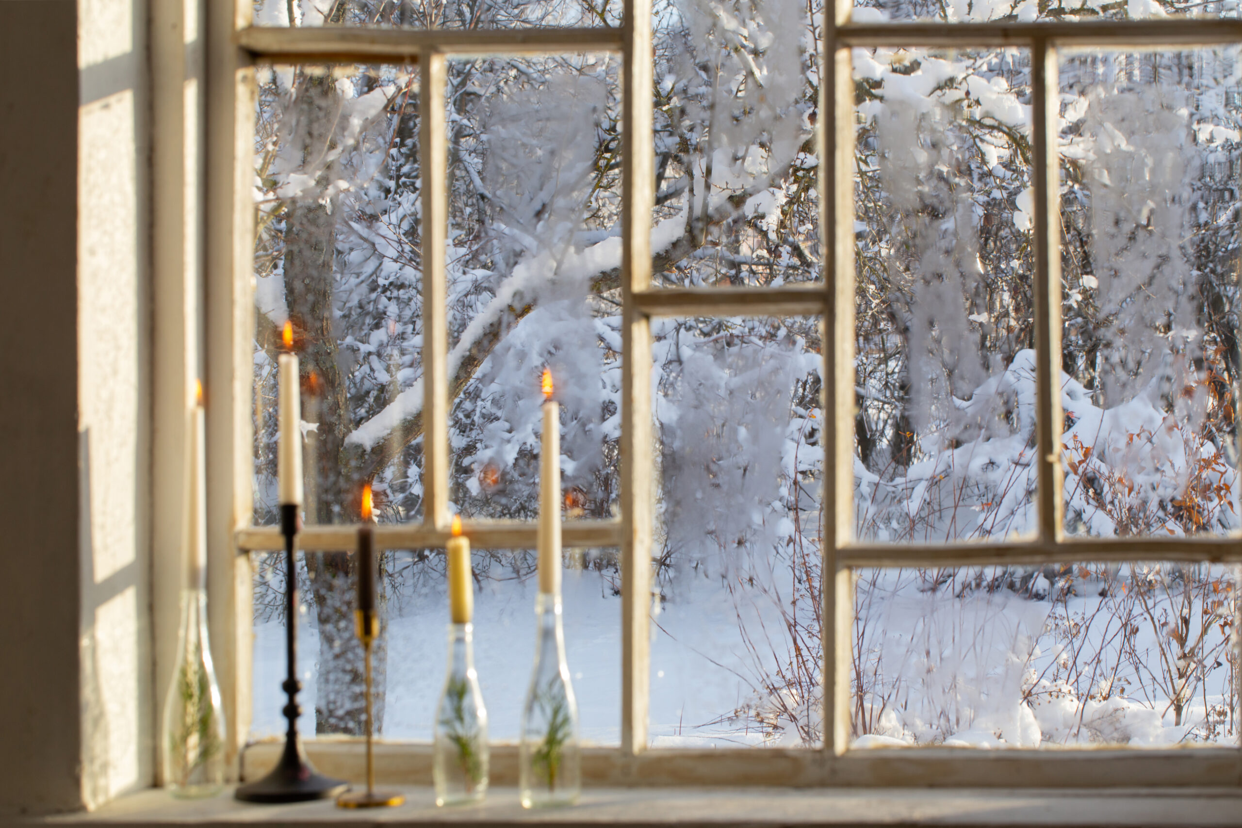 Старинное окно с видом на заснеженный сад, на подоконнике горящие свечи в подсвечниках и бутылках